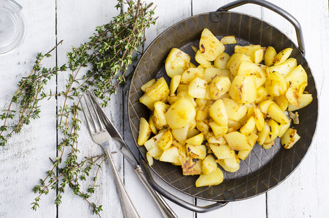 Gusseiserne Bratpfanne mit Bratkartoffeln, Thymian und Besteck auf Holz, lizenzfreies Stockfoto