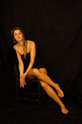 Porträt einer lächelnden Frau mit schwarzer Korsage, die auf einem Stuhl vor einem schwarzen Hintergrund sitzt - TCF004461
