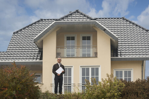 Immobilienmakler mit Unterlagen vor einem Wohnhaus stehend - PAF001112