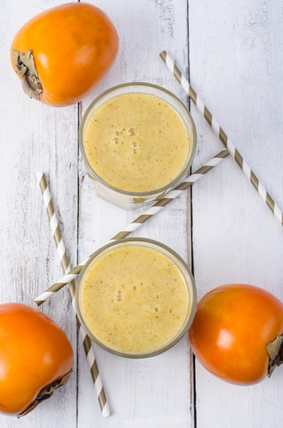 Zwei Gläser mit Kaki-Smoothie, Kakis und Trinkhalmen auf weißem Holz, lizenzfreies Stockfoto