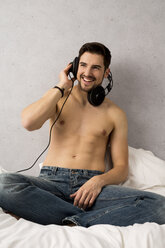 Mann ohne Hemd sitzt auf dem Bett und hört Musik mit Kopfhörern - SHKF000126