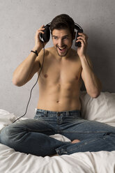 Mann ohne Hemd sitzt auf dem Bett und hört Musik mit Kopfhörern - SHKF000127