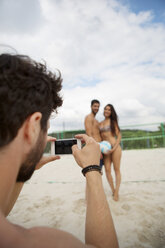 Young man taking cell phone picture of friends on beach volleyball field - WESTF020839