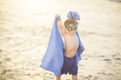 Kleiner Junge am Strand verkleidet als Superheld mit Maske und Handtuch, lizenzfreies Stockfoto