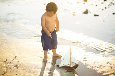 Boy on the beach playing with a toy wooden boat in the water - ZEF003429