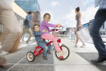 Mädchen und Junge fahren auf einem Dreirad zwischen einer Menschenmenge in einer Stadt - ZEF003968