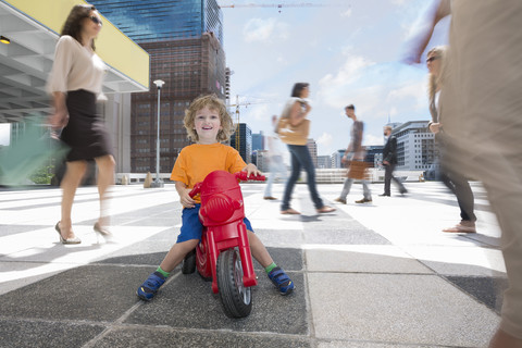 Junge fährt mit einem Plastikdreirad zwischen einer Menschenmenge in einer Stadt, lizenzfreies Stockfoto