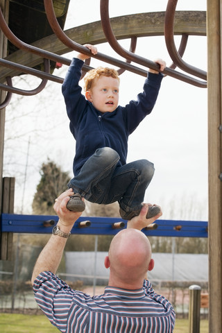 Vater hilft seinem Sohn auf dem Spielplatz beim Klettern auf dem Klettergerüst, lizenzfreies Stockfoto