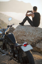 Südafrika, Kapstadt, Motorradfahrer sitzt auf einem Felsen an der Küste und fotografiert - ZEF003606