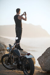 South Africa, Cape Town, motorcyclist standing on rock at the coast taking pictures - ZEF003608