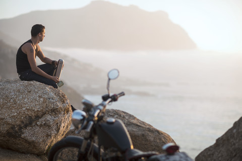 Südafrika, Kapstadt, Motorradfahrer sitzt auf einem Felsen an der Küste und genießt die Aussicht, lizenzfreies Stockfoto