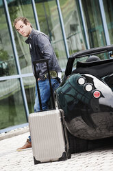 Young man with rolling suitcase leaning at his sports car - HHF005022