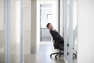 Businessman in suit sitting on chair leaning into hallway - ZEF003128