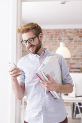 Smiling young man with cell phone and folders in office - WESTF020526