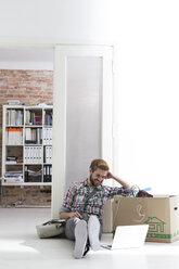 Young man sitting on floor in office using laptop next to cardboard box - WESTF020557