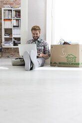 Junger Mann sitzt auf dem Boden im Büro und benutzt einen Laptop neben einem Karton - WESTF020558
