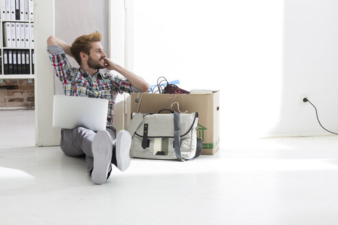 Junger Mann sitzt auf dem Boden im Büro und benutzt einen Laptop neben einem Karton - WESTF020559