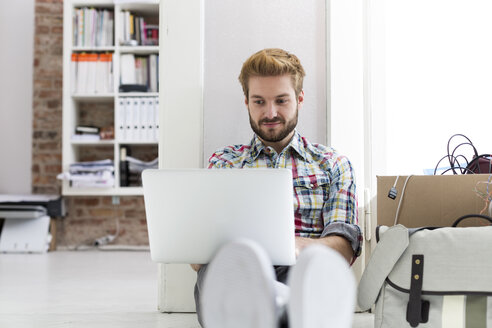 Junger Mann sitzt auf dem Boden im Büro und benutzt einen Laptop neben einem Karton - WESTF020560