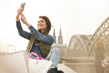 Germany, Cologne, smiling young woman with shopping bags taking a selfie - FEXF000259