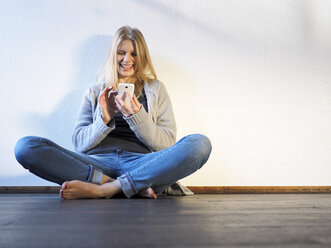 Happy young woman with smartphone sitting cross-leged on wooden floor - LAF001279