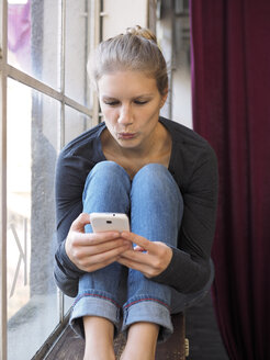 Junge Frau sitzt auf dem Fensterbrett und benutzt ihr Smartphone - LAF001284