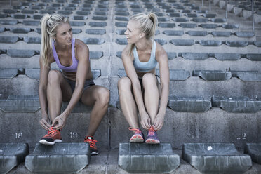 Two sportswomen sitting on grandstand of a stadium tying their shoes - ZEF004592