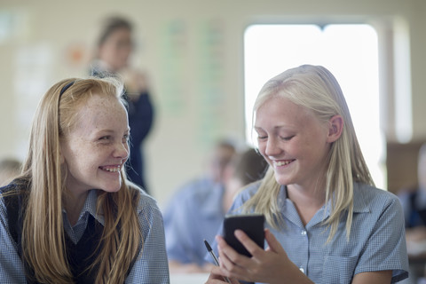 Zwei lächelnde Schulmädchen im Klassenzimmer mit Mobiltelefon, lizenzfreies Stockfoto