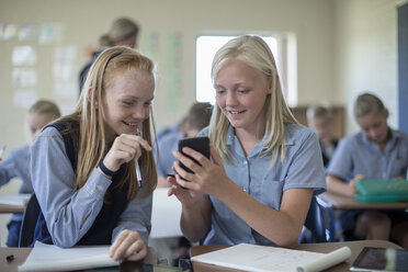 Zwei lächelnde Schulmädchen im Klassenzimmer schauen auf ihr Handy - ZEF004971