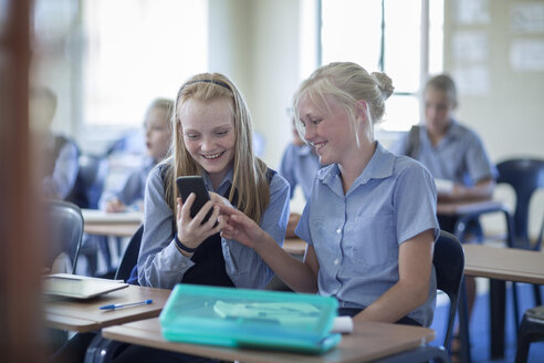 Zwei lächelnde Schulmädchen im Klassenzimmer schauen auf ihr Handy - ZEF004979
