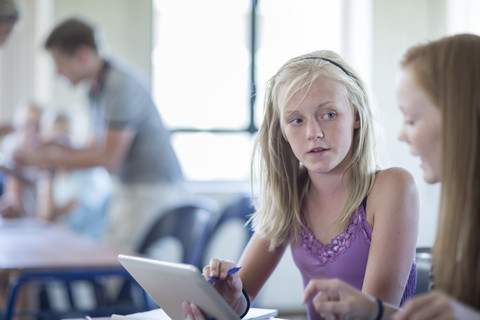 Zwei Schulmädchen im Klassenzimmer mit digitalem Tablet im Gespräch, lizenzfreies Stockfoto