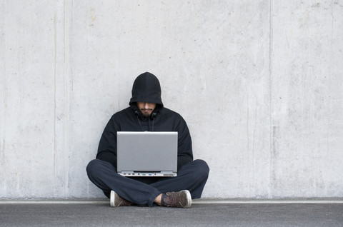 Hacker mit Laptop vor einer Betonwand sitzend, lizenzfreies Stockfoto