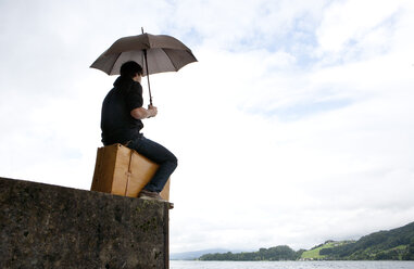 Junger Mann mit Regenschirm auf Koffer sitzend am Mondseeufer - WWF003680