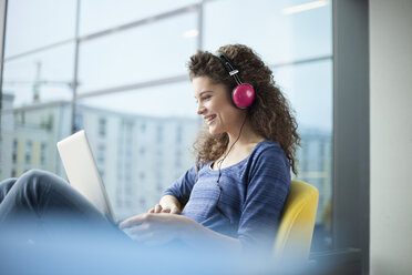 Lächelnde junge Frau mit Kopfhörern und einem Laptop am Fenster - RBF002326