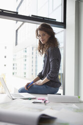 Porträt einer jungen Frau, die auf einem Schreibtisch im Büro sitzt - RBF002355