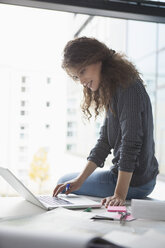 Junge Frau sitzt auf einem Schreibtisch im Büro und benutzt einen Laptop - RBF002356