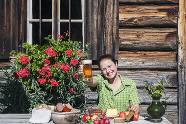 Smiling young woman sitting in front of Alpine cabin toasting with beer - HHF005085