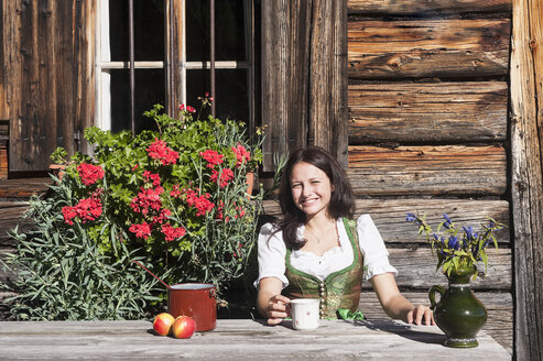 Smiling young woman sitting in front of Alpine cabin enjoying vacation - HHF005088