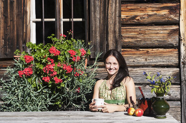 Smiling young woman sitting in front of Alpine cabin enjoying vacation - HHF005090