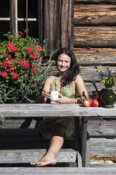 Smiling young woman sitting in front of Alpine cabin enjoying vacation - HHF005091