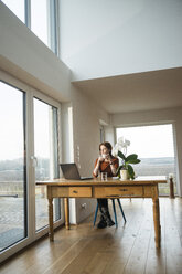 Young woman with laptop at wooden table looking out of window - UUF003203