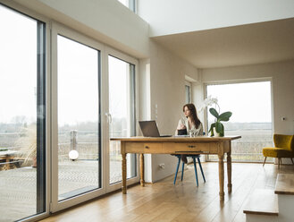 Junge Frau mit Laptop am Holztisch und Blick aus dem Fenster - UUF003205