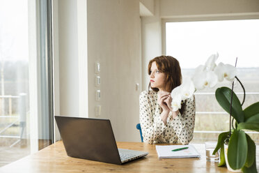 Junge Frau mit Laptop am Holztisch und Blick aus dem Fenster - UUF003209