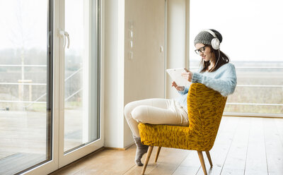 Smiling young woman sitting on chair with digital tablet and headphones - UUF003241