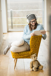 Smiling young woman sitting on chair with digital tablet and headphones - UUF003244