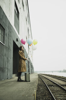 Junge Frau mit Koffer, die auf dem Bahnsteig einen Strauß Luftballons hält - UUF003262