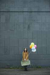Junge Frau mit Koffer, die einen Strauß Luftballons hält - UUF003276