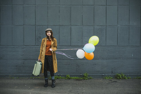 Junge Frau mit Koffer, die einen Strauß Luftballons hält - UUF003279