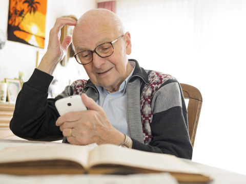 Alter Mann sitzt am Tisch und benutzt ein Mobiltelefon, lizenzfreies Stockfoto
