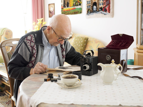 Alter Mann sitzt am Tisch und beschäftigt sich mit einer alten Kamera, lizenzfreies Stockfoto