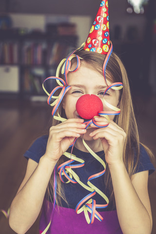 Kleines Mädchen mit Clownsnase und Mütze bläst Luftschlangen, lizenzfreies Stockfoto